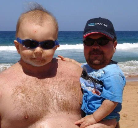 Manbabies — Shirtless man with baby's face holding baby with adult man's face at the beach