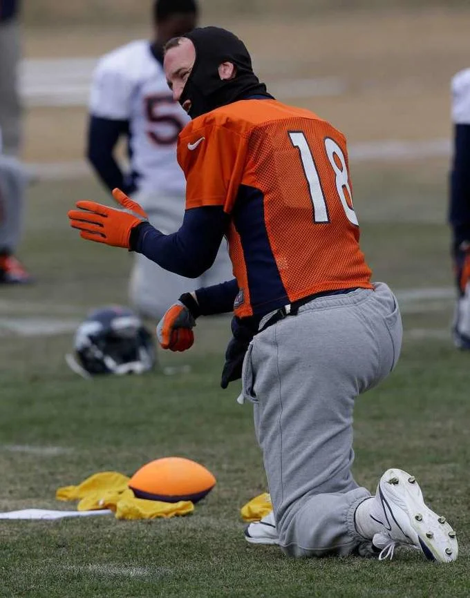 ManningFace — Peyton Manning kneeling in orange Broncos jersey number 18 wearing a black balaclava at practice