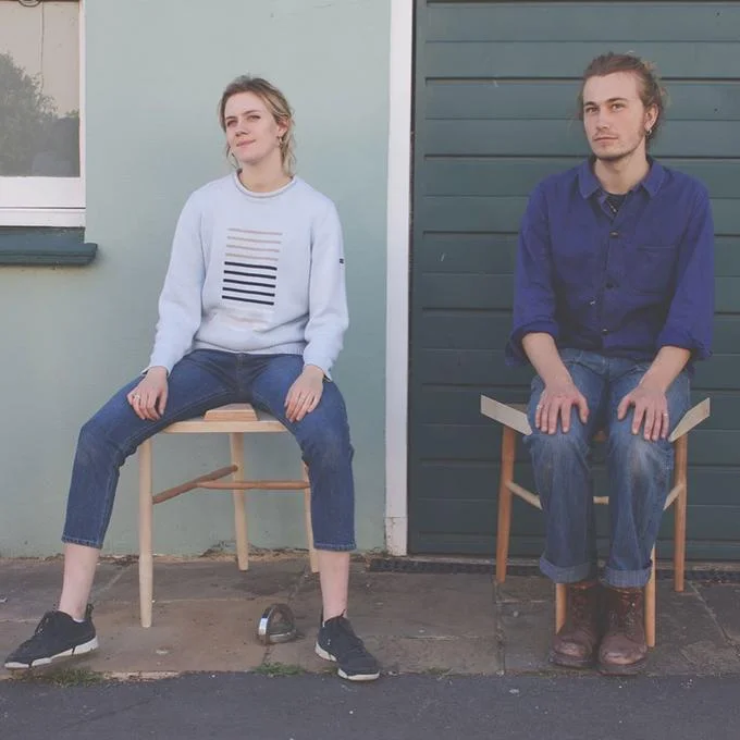 Two wooden anti-manspreading chairs with wedge-shaped seats against a blue wall