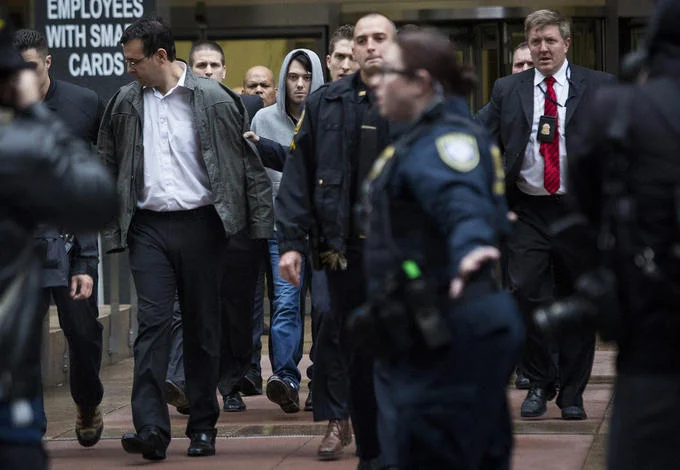 Martin Shkreli in a hoodie being led out by police officers after arrest