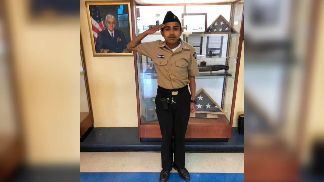 Me When X Tell Me to Do Something — Young JROTC cadet in tan uniform saluting beside a glass case with folded flags and a framed officer portrait.