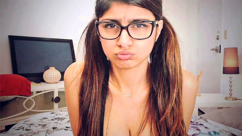 Mia Khalifa in black-framed glasses making a pout face in a bedroom