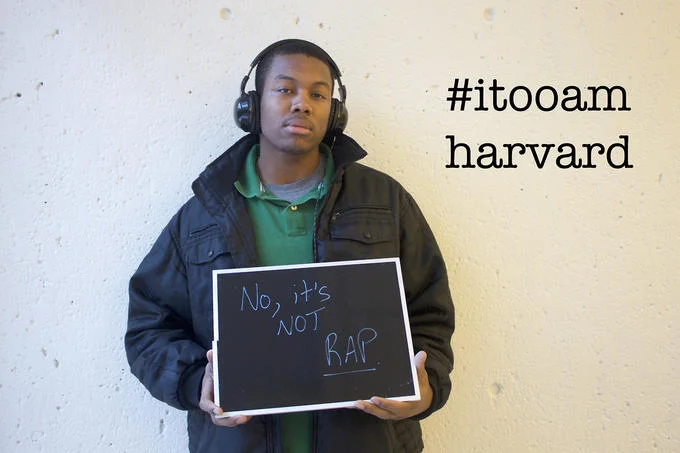 Microaggression — Black student with headphones holding sign 'No, it's NOT RAP' with #itooamharvard hashtag