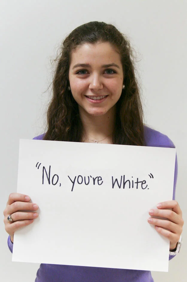 Microaggression — Smiling young woman in purple shirt holding sign 'No, you're white.'