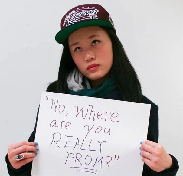 Microaggression — Asian woman in cap holding sign saying 'No, where are you REALLY from?'