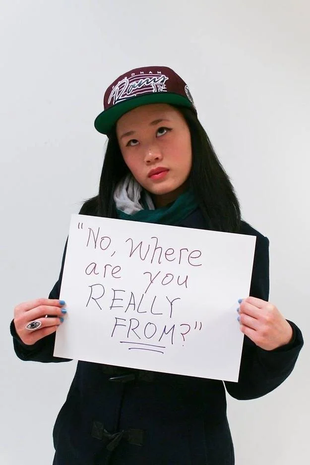 Microaggression — Full body shot of Asian woman holding 'No, where are you REALLY from?' sign