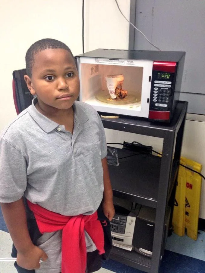 Minor Mistake Marvin — Portrait of young boy standing beside microwave with neutral, slightly regretful facial expression.