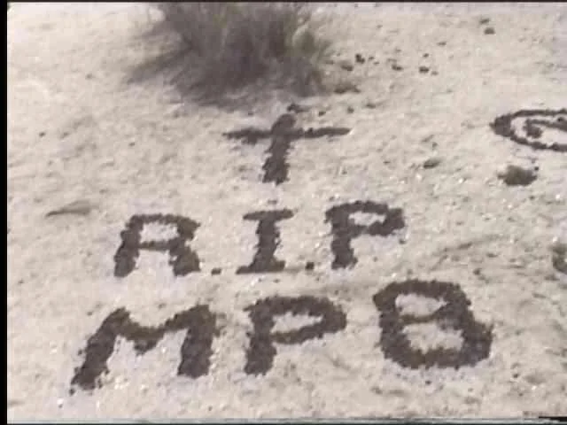 Mojave Phone Booth — Grainy black-and-white photograph showing 'R.I.P. MPB' inscription with cross symbol on desert floor.