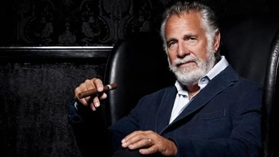 The Most Interesting Man in the World — Distinguished older man with gray beard in dark pinstriped suit seated with raised hand gesture.