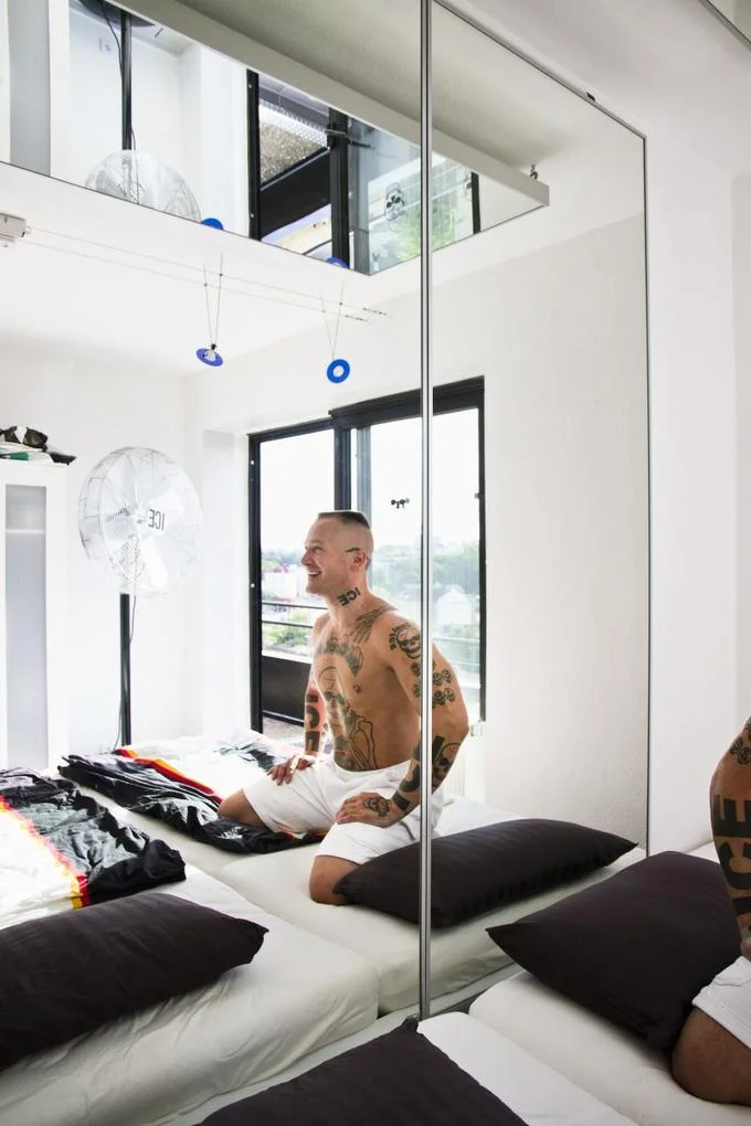 Mr. Cool Ice — Tattooed man laughing on a bed in a minimalist apartment with an ICE-branded fan nearby