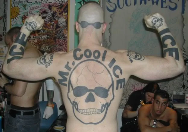 Wide back view of Mr. Cool Ice flexing his arms to display the skull back tattoo inside a tattoo parlor