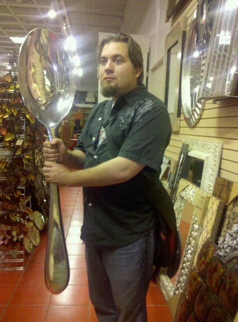 My Spoon Is Too Big — Man holding a giant decorative spoon in a home decor store