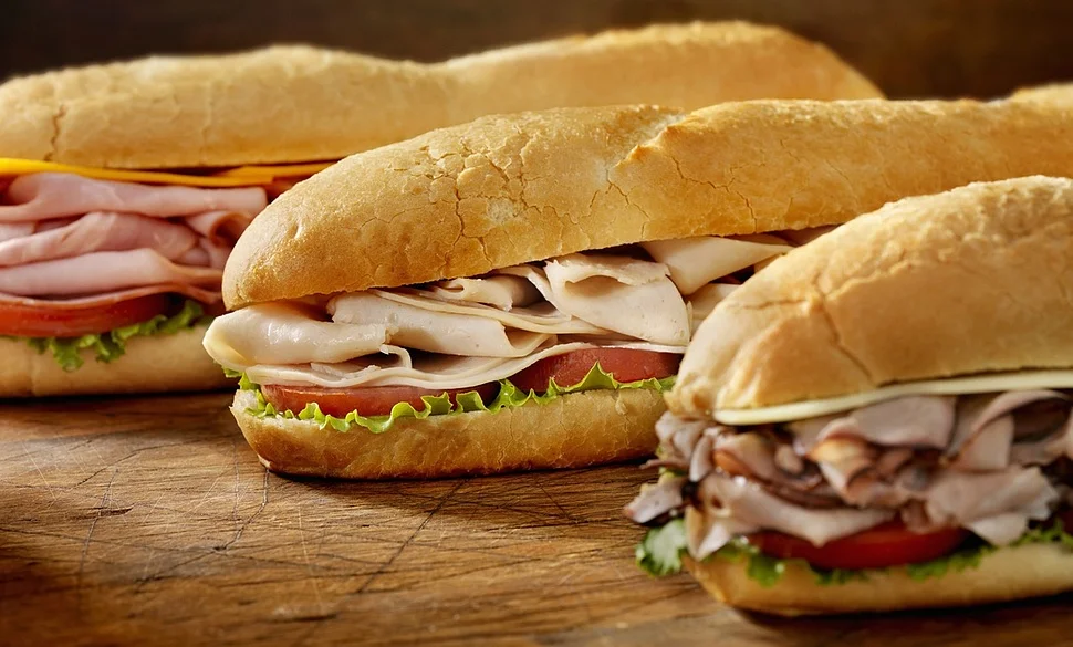 My Subs Have Unionized — Three submarine sandwiches with various meats and vegetables on a wooden surface