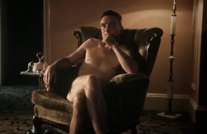 Naked Oppenheimer In A Chair — Cillian Murphy as Oppenheimer sits naked in upholstered chair with hand near mouth.