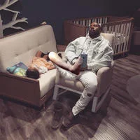 Napping Kanye — Kanye West naps on a display couch in light-colored clothing while his daughter North West rests on a smaller couch beside him.
