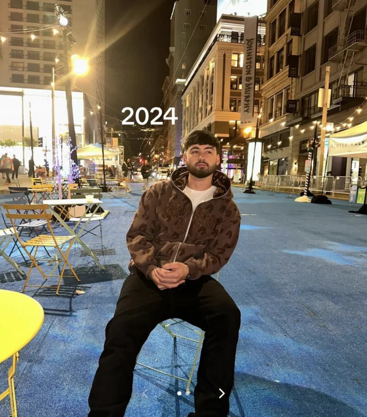 Noah Boat N Em — Grown-up Noah with a beard in a brown hoodie sitting outdoors at night in 2024.
