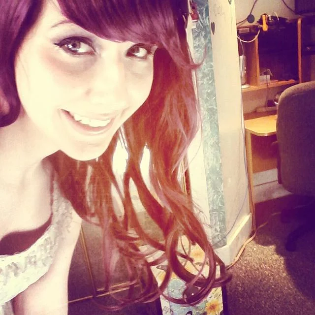 Smiling selfie of Nyanners with long reddish-purple hair and dark eye makeup in a bedroom setting.