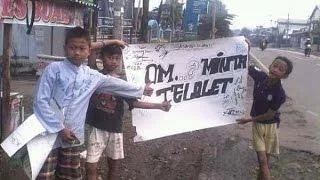 Indonesian youths holding handwritten 'OM TELOLET OM' sign on roadside street.