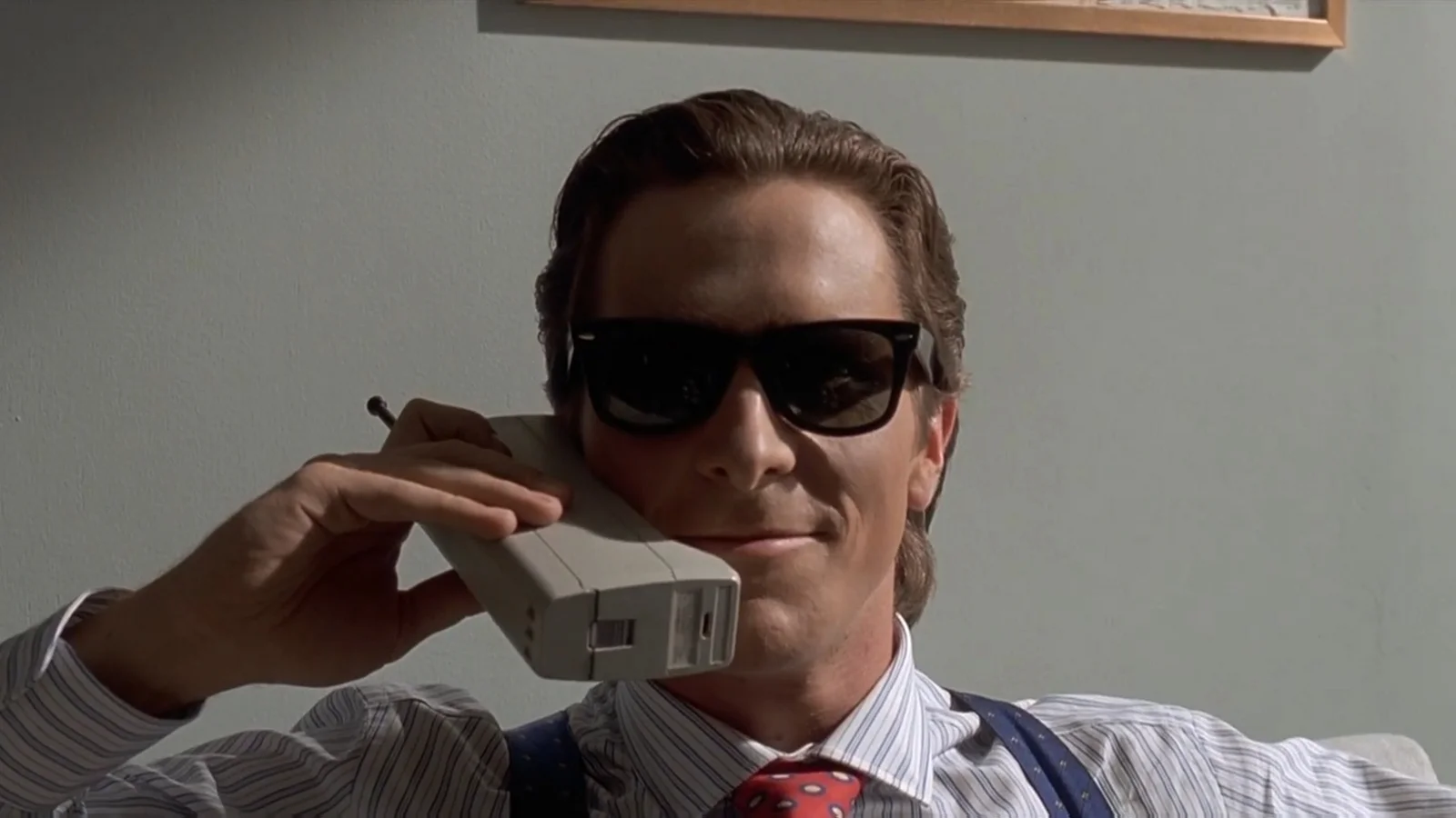 Patrick Bateman wearing sunglasses and holding a business card, embodying obsession with status symbols.