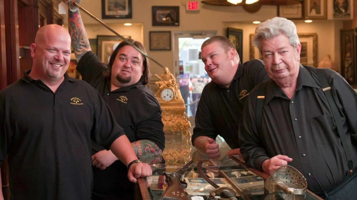 The four main cast members of Pawn Stars posing in the Gold & Silver Pawn Shop with antiques on display.