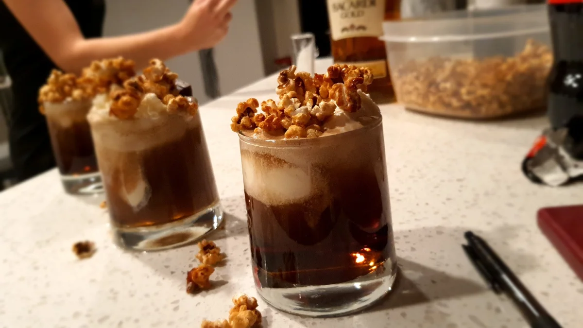 Pissy Shitties — Three glasses of a dark cocktail topped with caramel popcorn and cream on a kitchen counter.