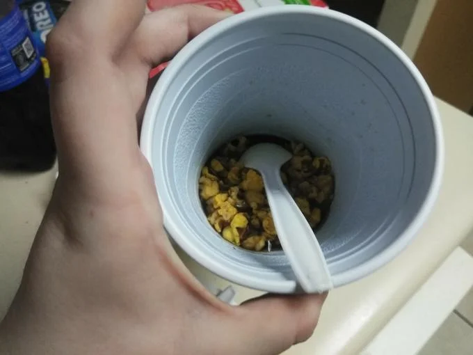 Pissy Shitties — Plastic cup of popcorn soaked in cola with a spoon inside, held by a hand.