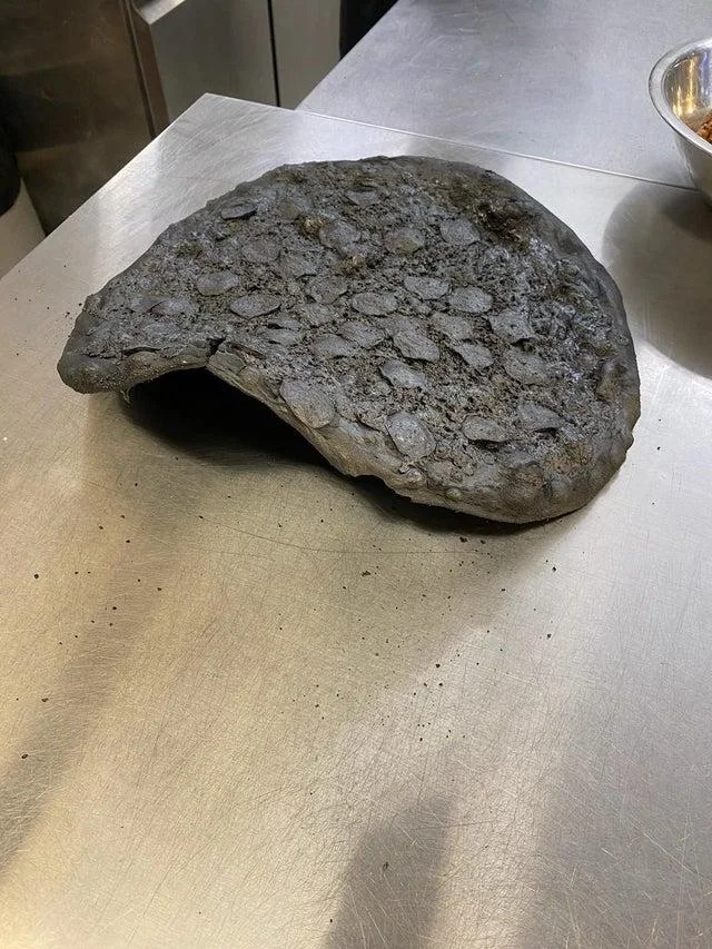 Pizza Crimes — A deeply charred and burnt pizza that has folded over itself showing severe overcooking and discoloration.