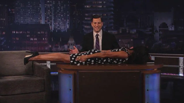 A late-night television host sits at his desk while someone lies horizontally in perfect planking position on the desk behind him.