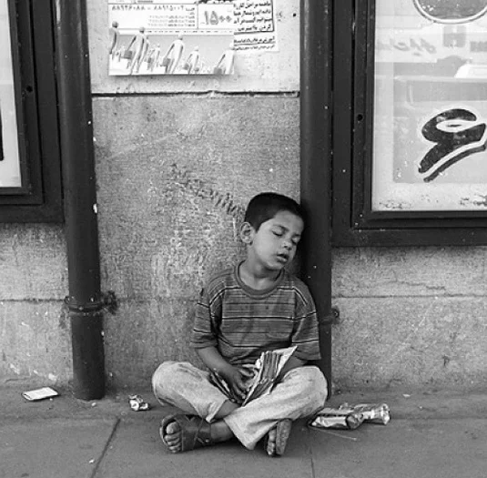 Poor Kids Of Tehran — A black and white photograph of a young boy sitting against a wall with head drooping and eyes closed near storefronts.