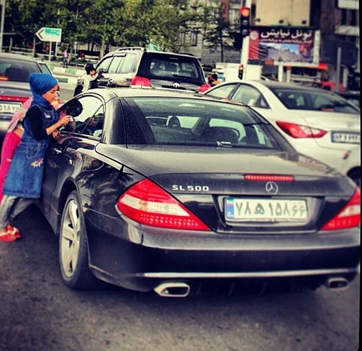 Poor Kids Of Tehran — A small child in blue approaching a luxury Mercedes-Benz SL500 at a Tehran traffic light window.