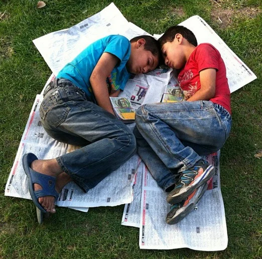 Poor Kids Of Tehran — Two young boys asleep on makeshift beds of spread newspapers on a patch of grass.