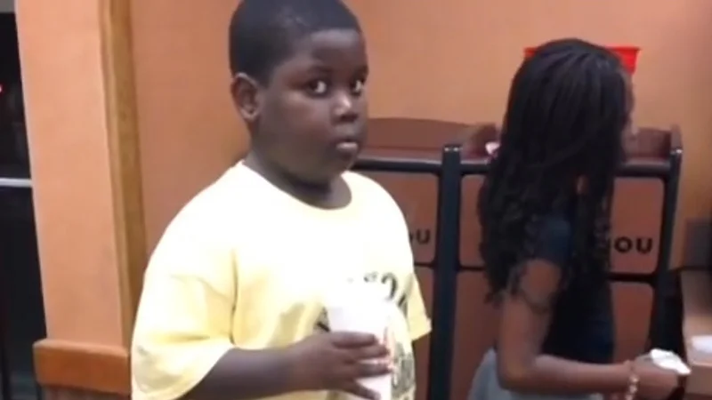 Popeyes Kid — Young Black boy in yellow shirt holding a cup at Popeyes with wide-eyed surprised expression next to a woman