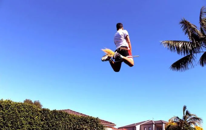Pottering — Person leaping into a blue sky above rooftops, straddling a broomstick