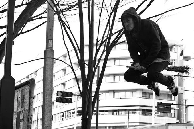 Black and white photo of a hooded person pottering on a broom in a Japanese city street