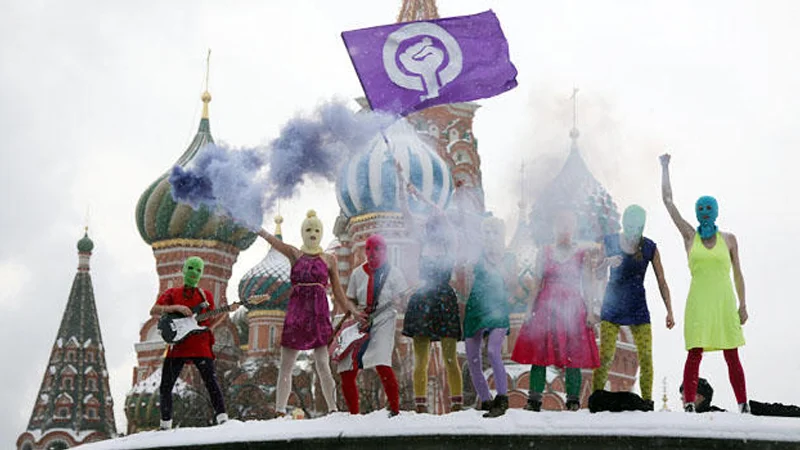 Pussy Riot members in colorful balaclavas performing on Red Square in front of Saint Basil's Cathedral with purple smoke and a flag.