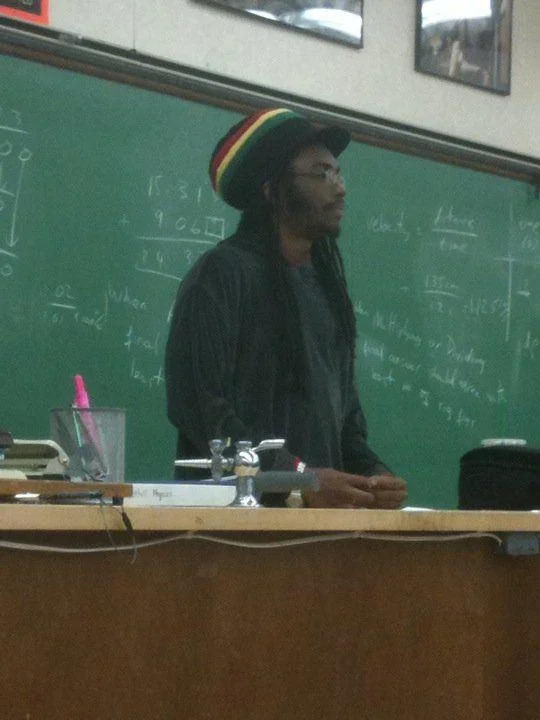 Rasta Science Teacher — Physics teacher wearing a rasta beanie and dreadlocks in front of a chalkboard covered in equations