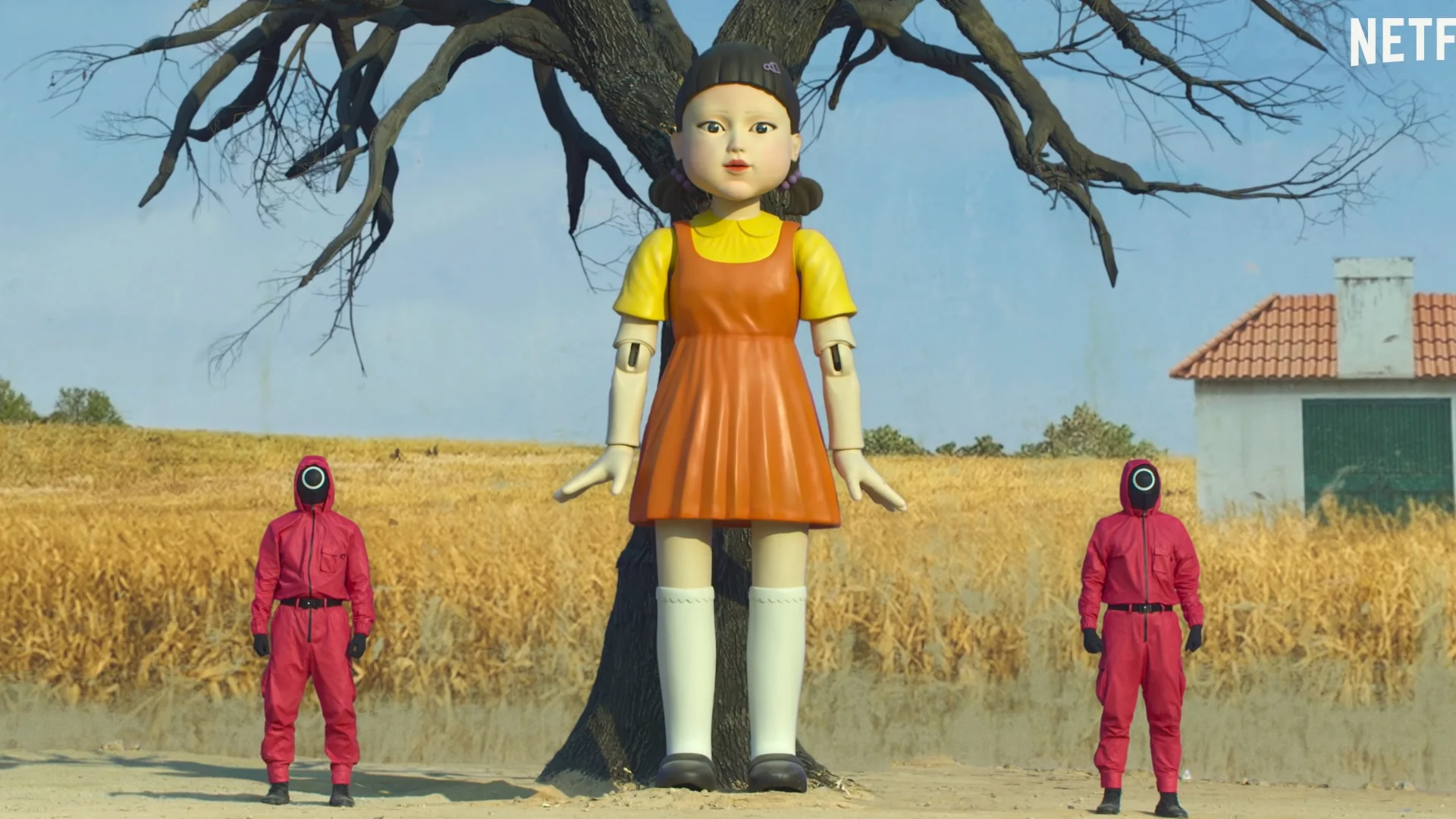 Red Light, Green Light — Giant Squid Game doll in orange dress standing between two red-suited guards in front of a tree
