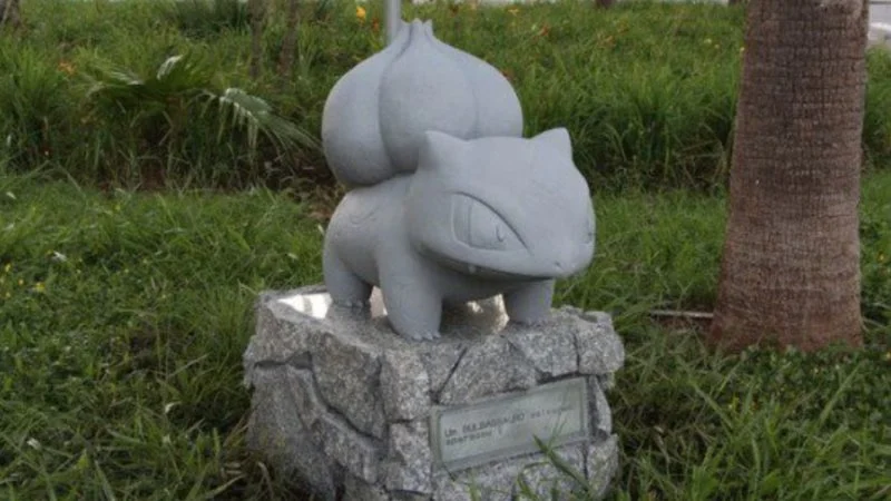 Replace Racist Statues — A clean outdoor photograph of a stone or granite Bulbasaur Pokémon statue on a rock plinth set in green grass with palm tree in