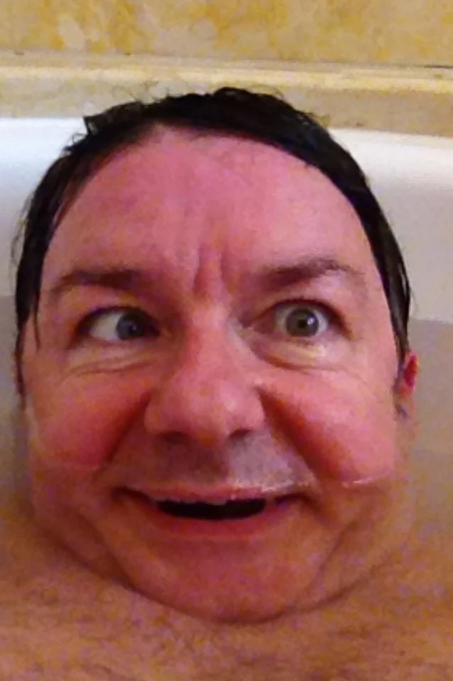 Ricky Gervais in a bathtub with wet hair pulling a wide-eyed silly face.