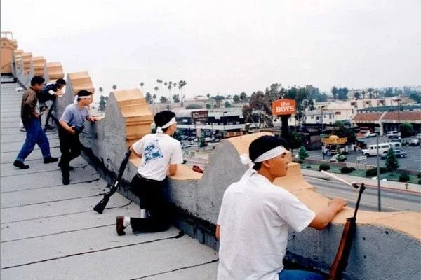 Roof Koreans — A historic 1992 photograph of several Korean-American men wearing white bandanas, crouching with rifles behind rooftop parapet wall overlooking