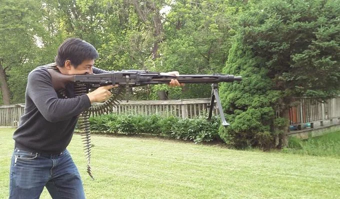 Roof Koreans — A modern photograph of an Asian man in green suburban backyard firing a large belt-fed machine gun at recreational shooting event.