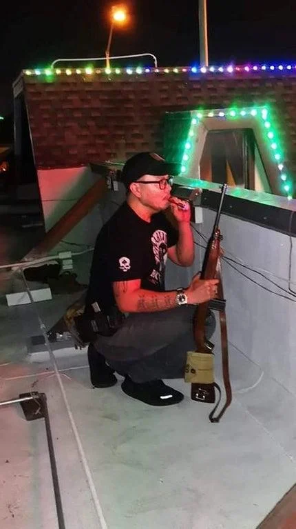 Roof Koreans — A nighttime photograph of man crouching on rooftop holding rifle with colorful holiday lights decorating nearby structure.