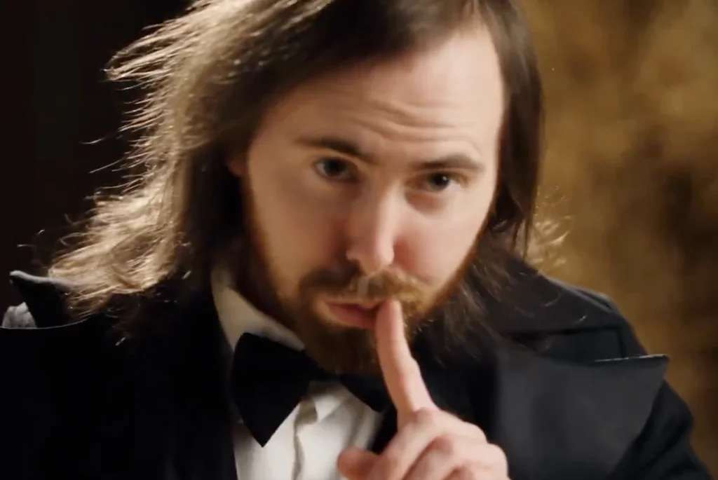 Close-up of a long-haired, bearded man in a black tuxedo and bow tie pressing a finger to his lips as Rumpelstiltskin.