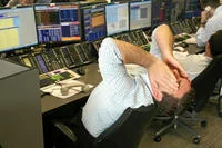 Sad Guys On Trading Floors — A trader at multi-screen financial workstation leaning far back in chair with both hands interlocked behind head in stressed