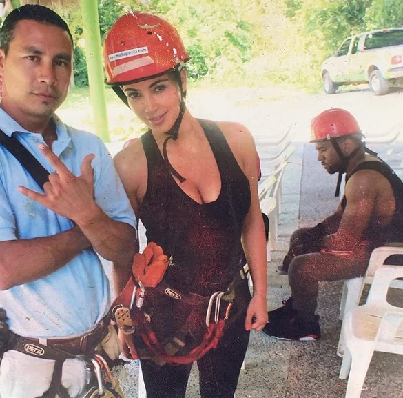 Sad Kanye — Kim Kardashian posing with guide while Kanye West sits slumped on a plastic chair in the background wearing a red helmet
