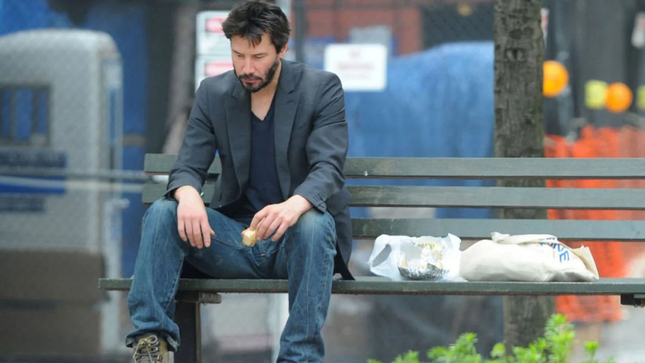 Sad Keanu — Keanu Reeves sitting alone on a park bench eating a sandwich, looking downcast.