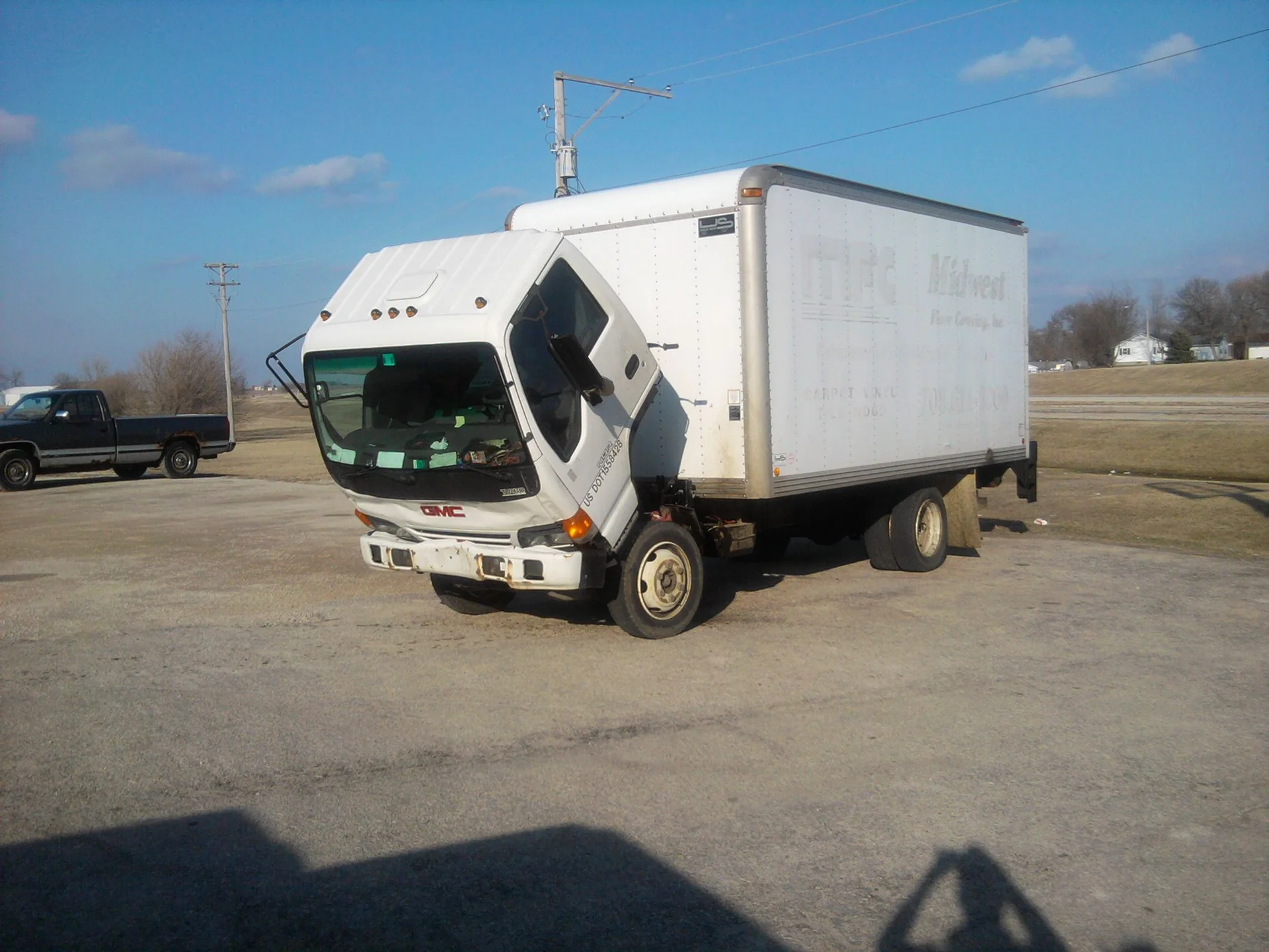 Sad Truck — White GMC box truck with tilted forward cab that appears to look sad, parked on pavement under a blue sky.