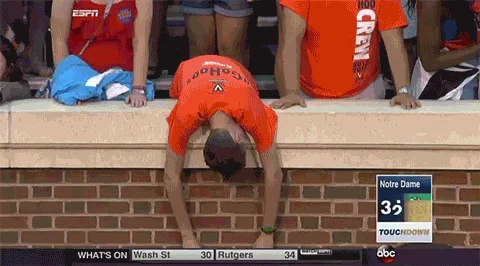 Sad Virginia Fan — A Virginia Cavaliers fan in orange UVA shirt bends over brick stadium wall in despair while ESPN broadcast shows 'Notre Dame 32 TOUCHDOWN'