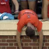 A tight close-up shot of Sad Virginia Fan in bright orange UVA shirt bent entirely over stadium brick wall in complete defeat with ESPN broadcast bug faintly