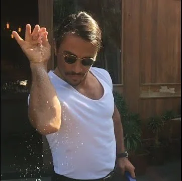 Salt Bae — The original viral photo of Turkish chef Nusret Gökçe mid-sprinkle with raised arm, white sleeveless shirt, and round sunglasses.